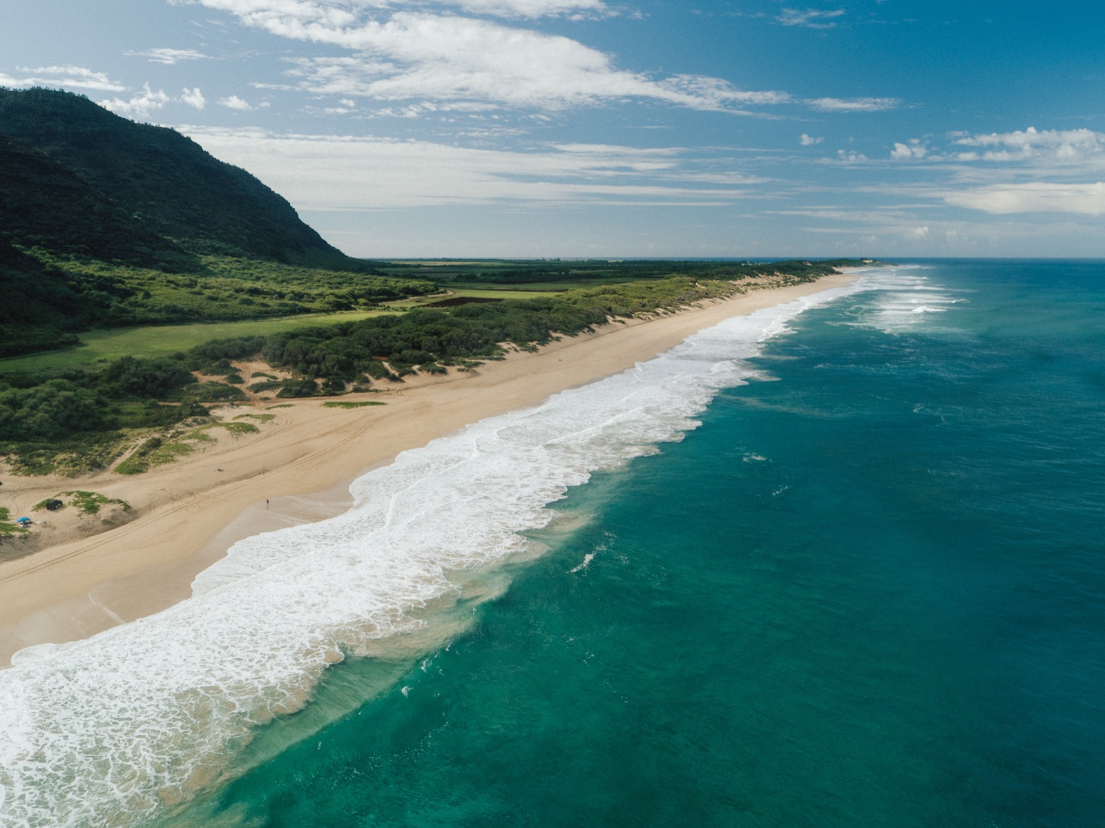 Aerial view of a long sandy beach on the West Kauaʻi coastline, with surf rolling in and green hills behind