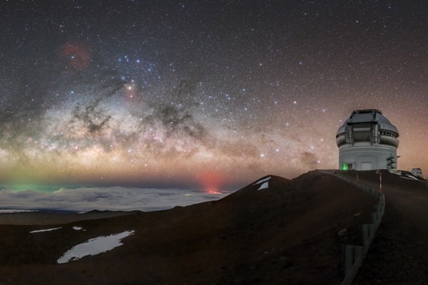 The Gemini North observatory on Maunakea at night under the Milky Way, above a sea of clouds, with the red glow of Kīlauea's eruption in the distance