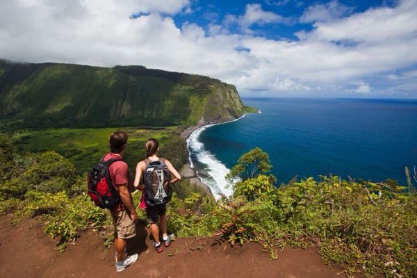 Two hikers at the Waipiʻo Valley lookout above the black-sand bay and sea cliffs