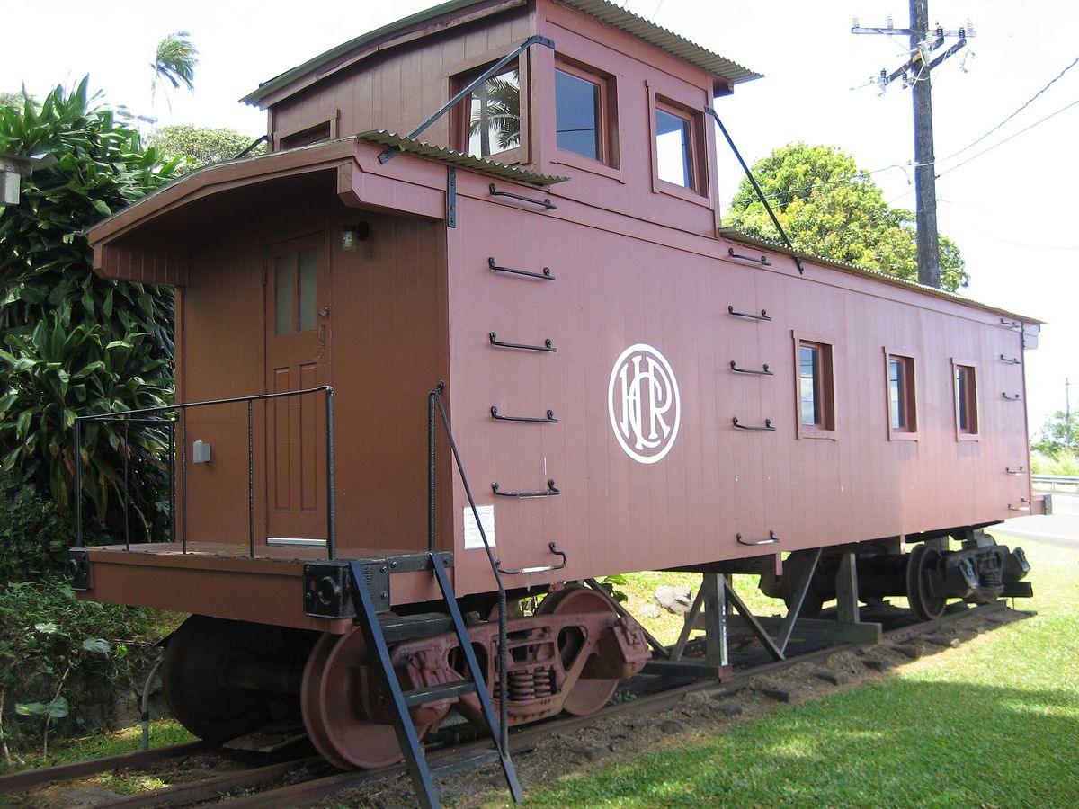 A restored maroon wooden railroad caboose displayed on a short track beside tropical plants.