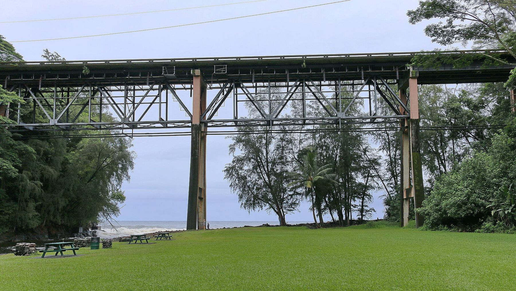 The tall steel trestle of the Highway 19 bridge spanning Kolekole Beach Park, with lawn and picnic tables below and the ocean beyond.
