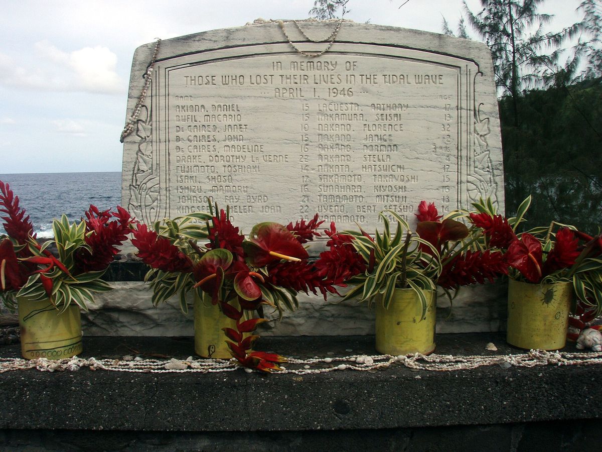 A stone memorial inscribed ‘In memory of those who lost their lives in the tidal wave, April 1, 1946’, with red flower offerings along its base.