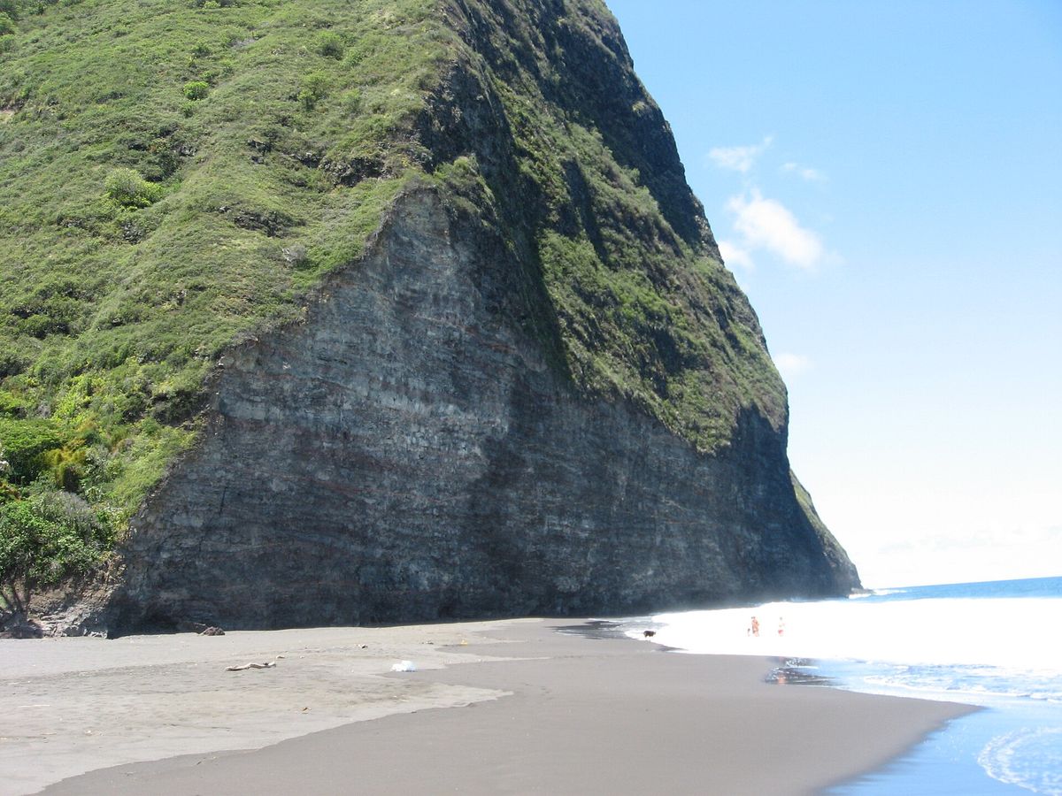Black sand beach at the mouth of Waipiʻo Valley beneath a towering green and grey sea cliff, with waves washing in.