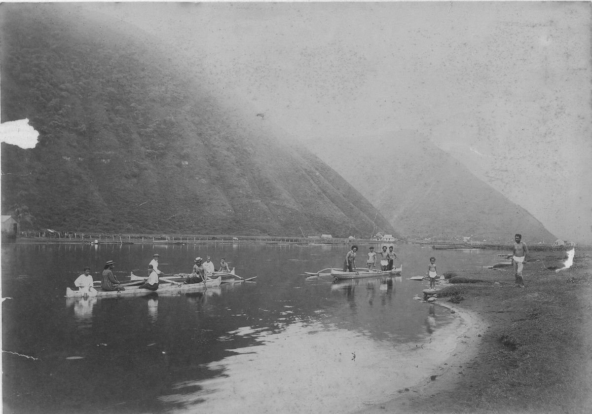 Historic black-and-white photograph of people in outrigger canoes on the calm stream at the mouth of Waipiʻo Valley, with the valley wall rising behind.
