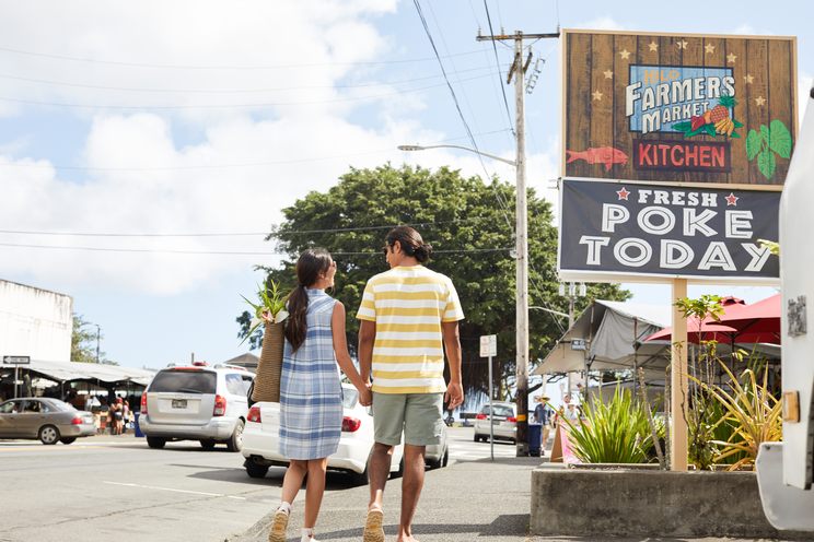 A couple walking toward the Hilo Farmers Market past a sign reading Farmers Market Kitchen, Fresh Poke Today
