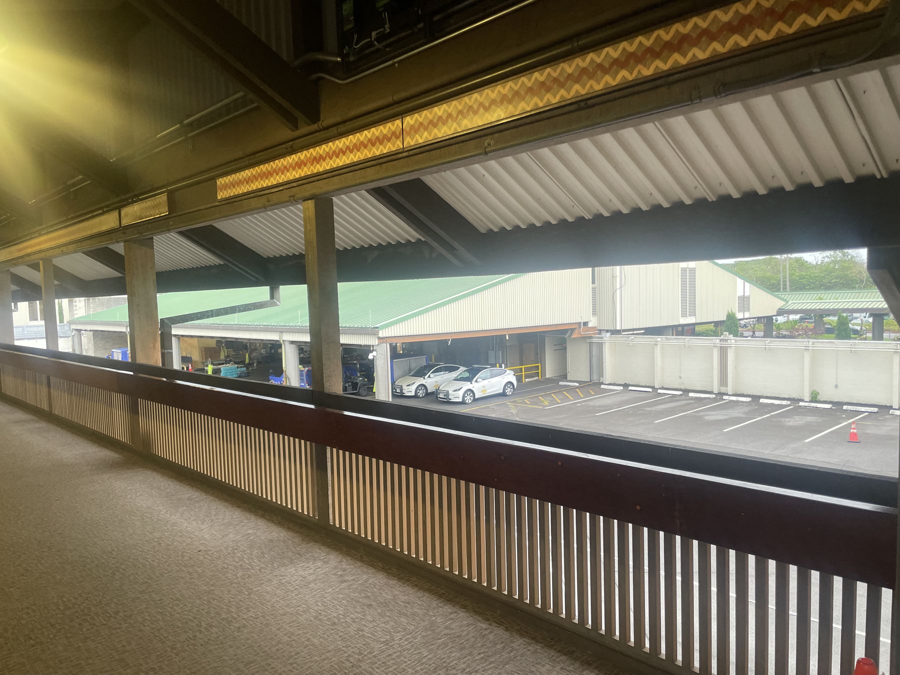 Covered open-air walkway at Hilo International Airport with wooden railings, looking out to a parking lot and green hills