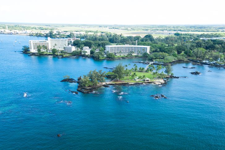 Hotels along the shore of Hilo Bay seen from above, with the small wooded island of Moku Ola in the foreground