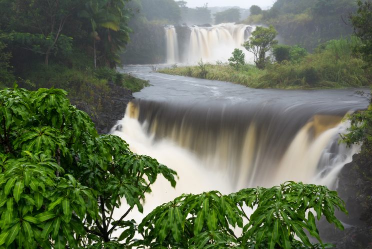 A wide river tumbling over several tiers of rock, blurred white by a long exposure, with green foliage in front