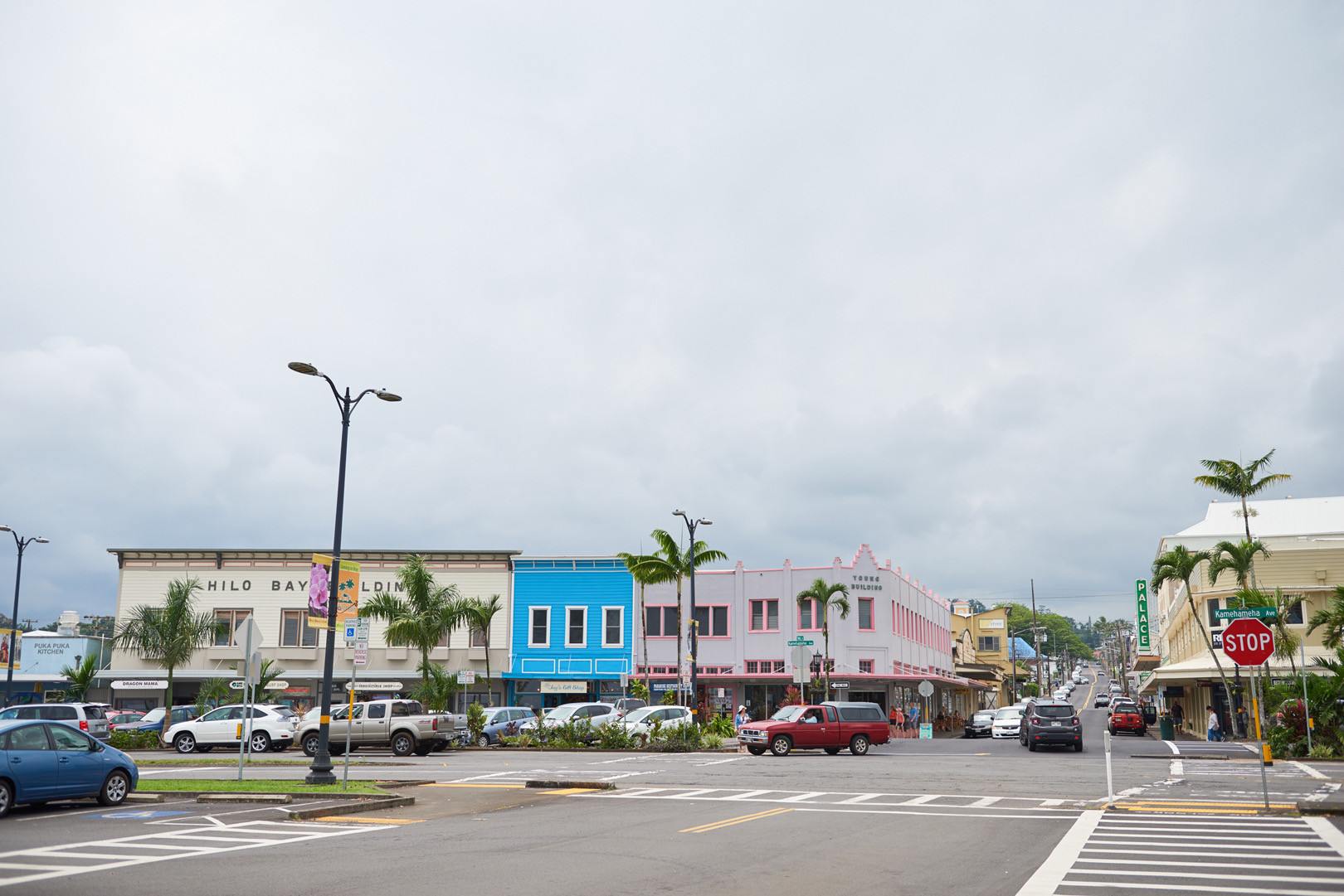 A downtown Hilo intersection under a cloudy sky, with colorful two-story storefronts, palm trees and parked cars along the street