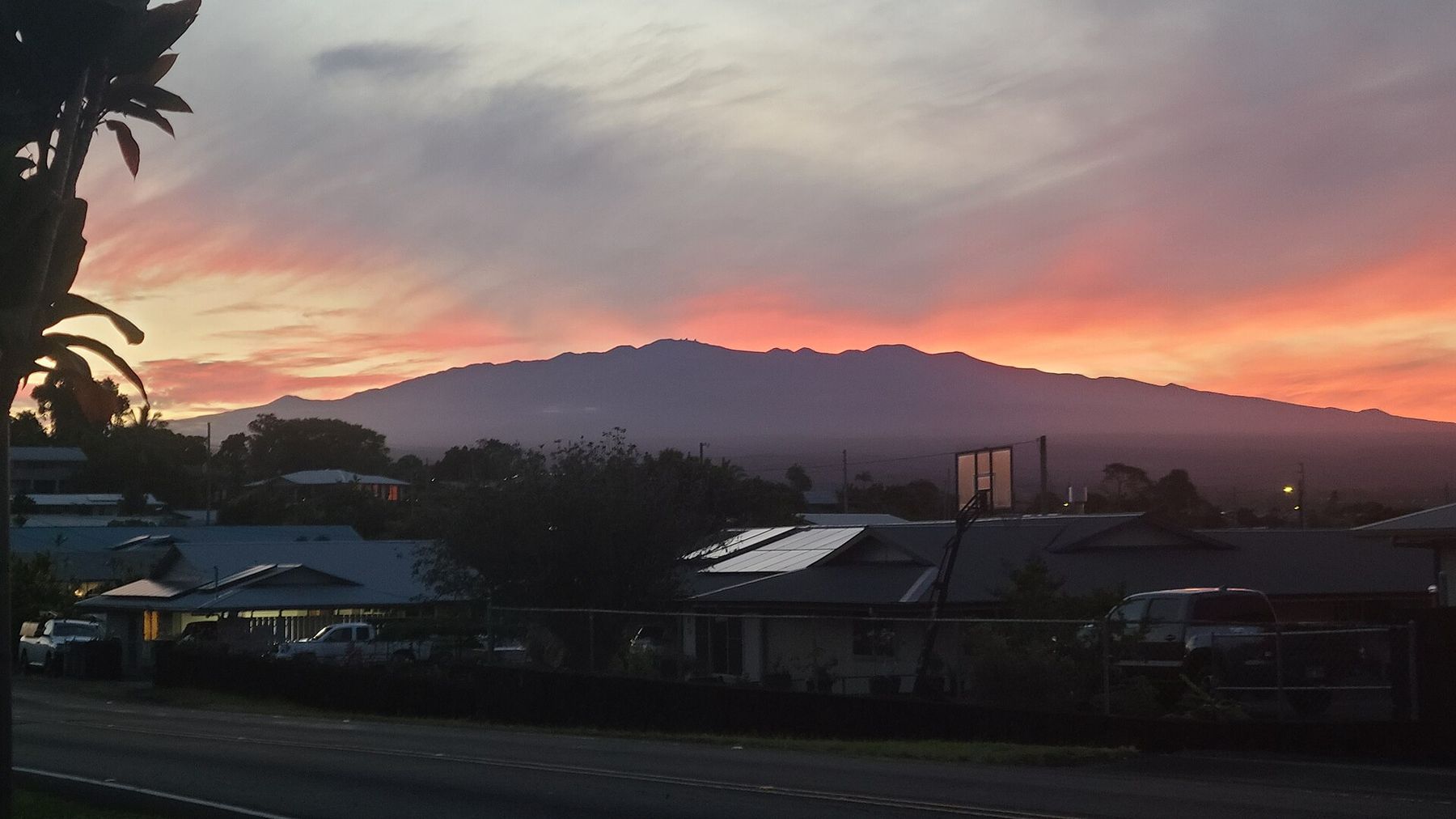 Maunakea silhouetted against an orange and pink sunset sky above Hilo rooftops