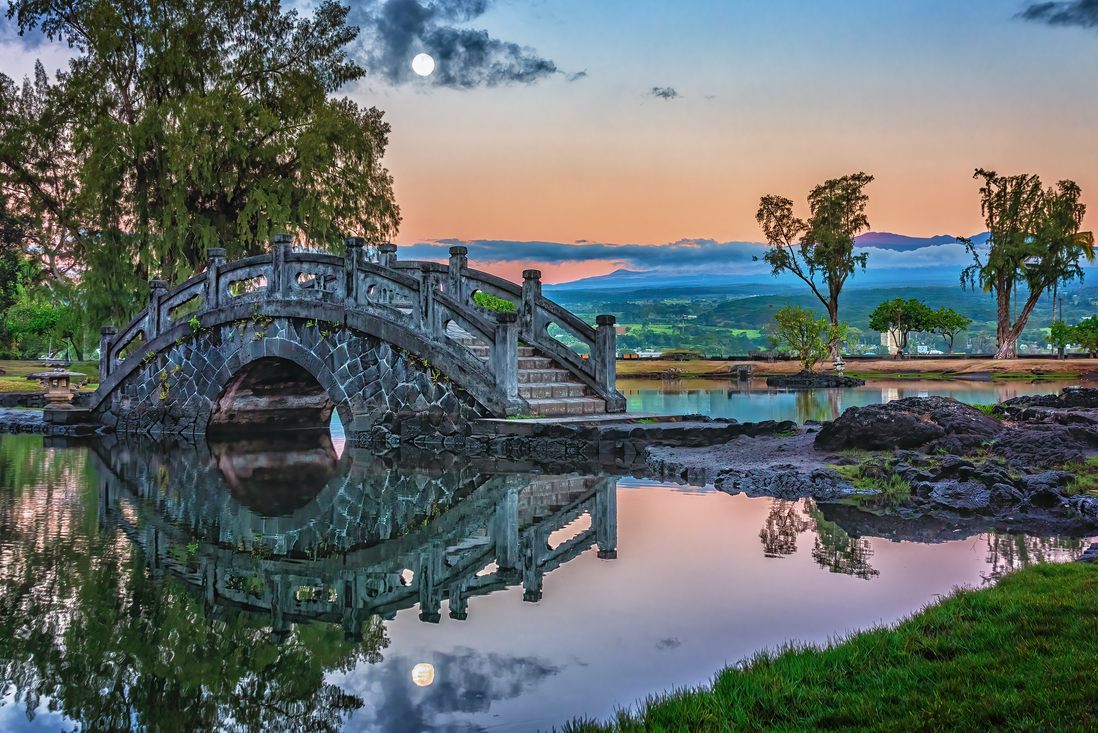 Arched bridge and trees reflected in a pond at Liliʻuokalani Gardens under a pink and blue sunset sky