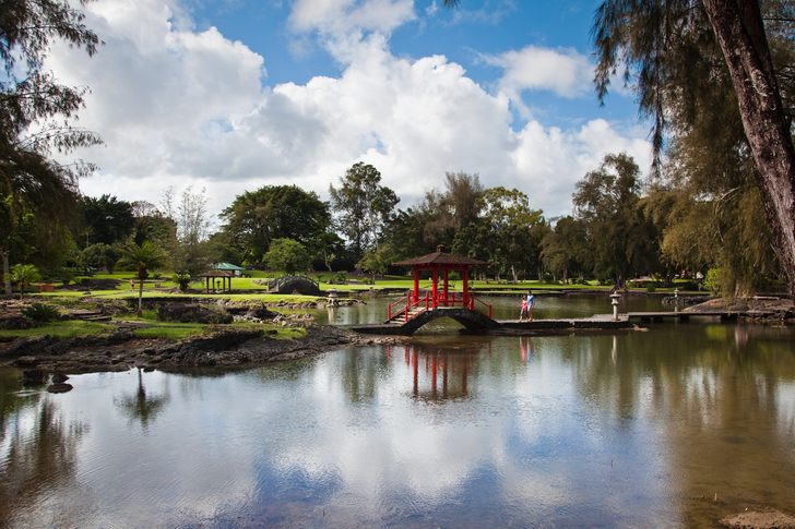 A small red bridge across a reflecting pond in Liliʻuokalani Gardens under white clouds