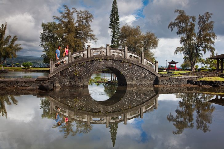 Visitors on an arched stone bridge reflected in a pond at Liliʻuokalani Gardens