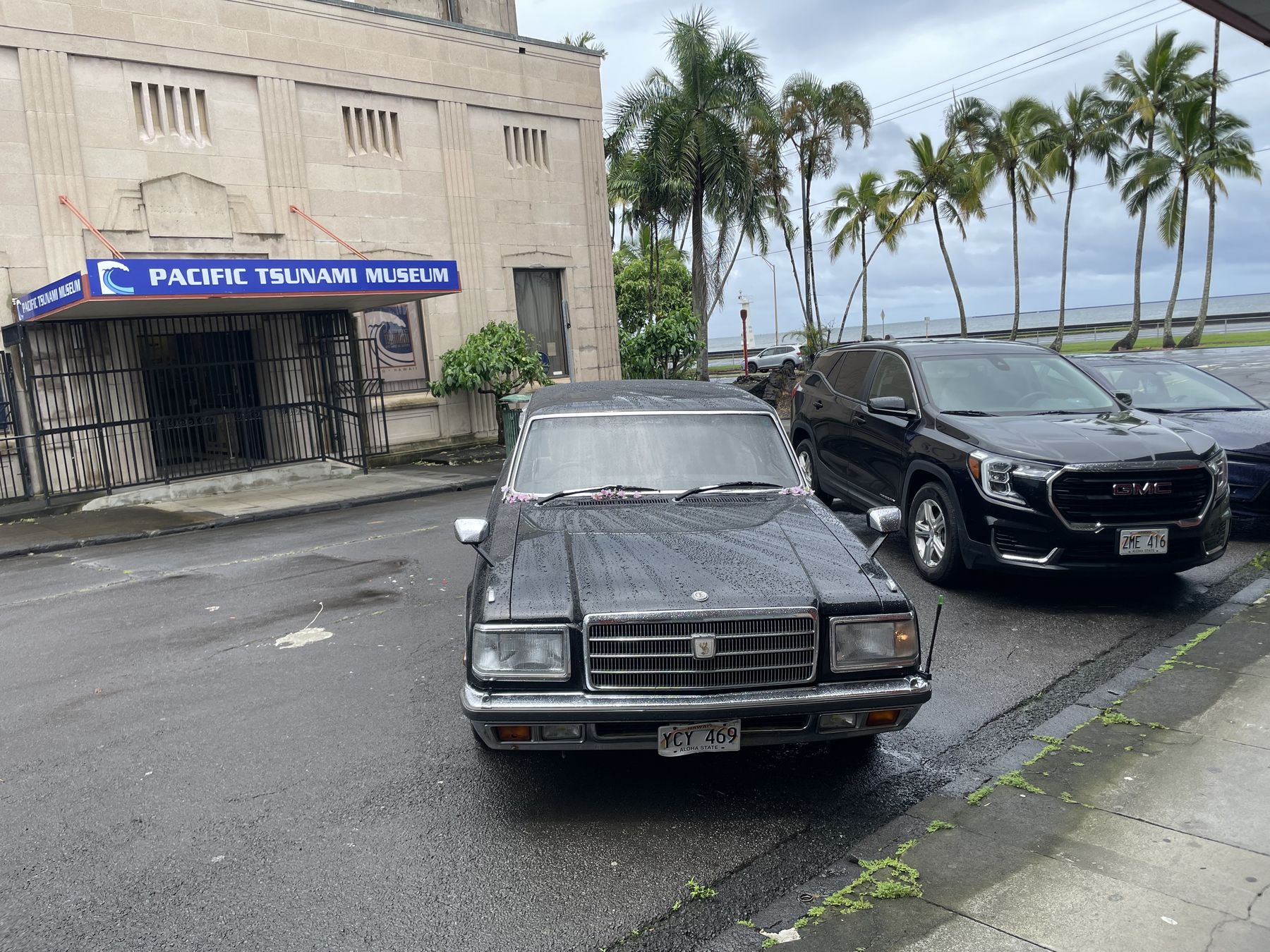 The stone facade of the Pacific Tsunami Museum with its blue sign, cars parked out front, and palm trees behind