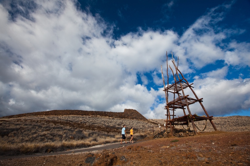 Visitors walking toward Puʻukoholā Heiau across dry ground, with a tall wooden anuʻu tower beside the path