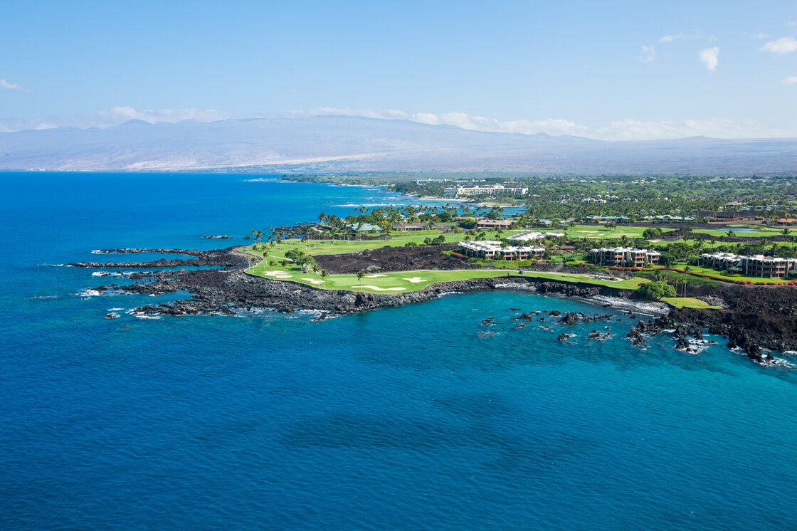 Aerial view of the Kohala Coast: vacation rentals and a golf course on a black lava point above deep blue water, with mountains on the horizon