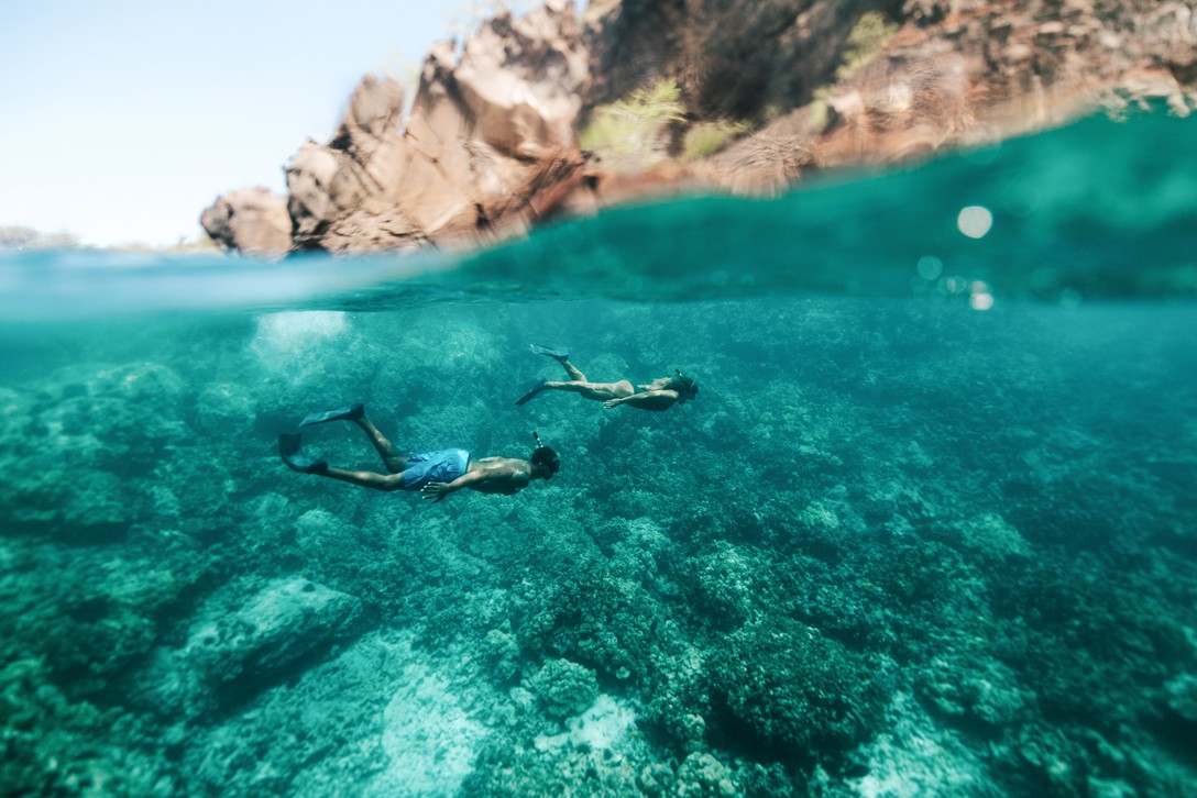 Two snorkelers swimming over coral in clear turquoise water below rocky cliffs at Kealakekua