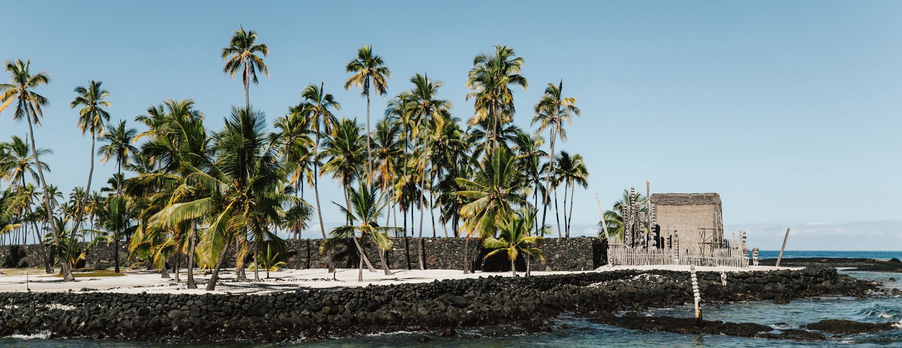 Coconut palms, lava rock walls and the thatched Hale o Keawe with carved kiʻi at Puʻuhonua o Hōnaunau