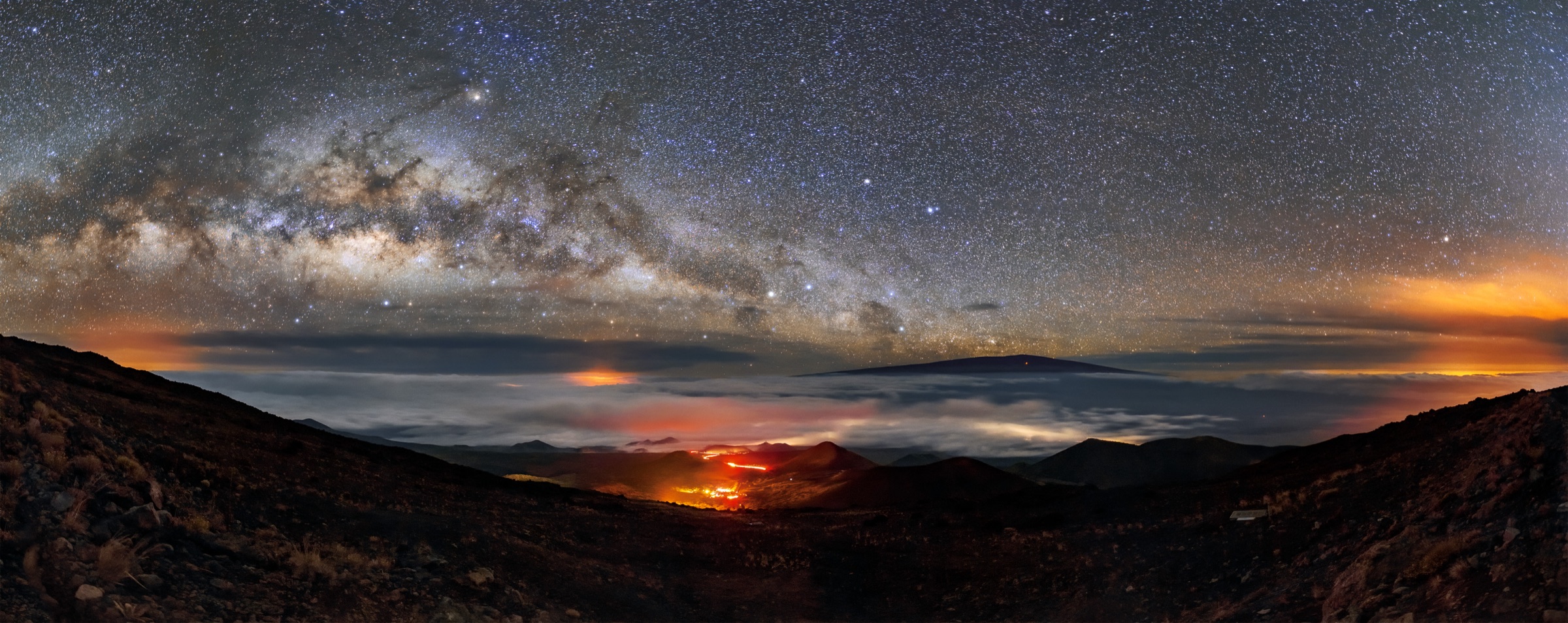 The Milky Way arching over the slopes of Maunakea, with Mauna Loa rising above a sea of clouds and town lights glowing below