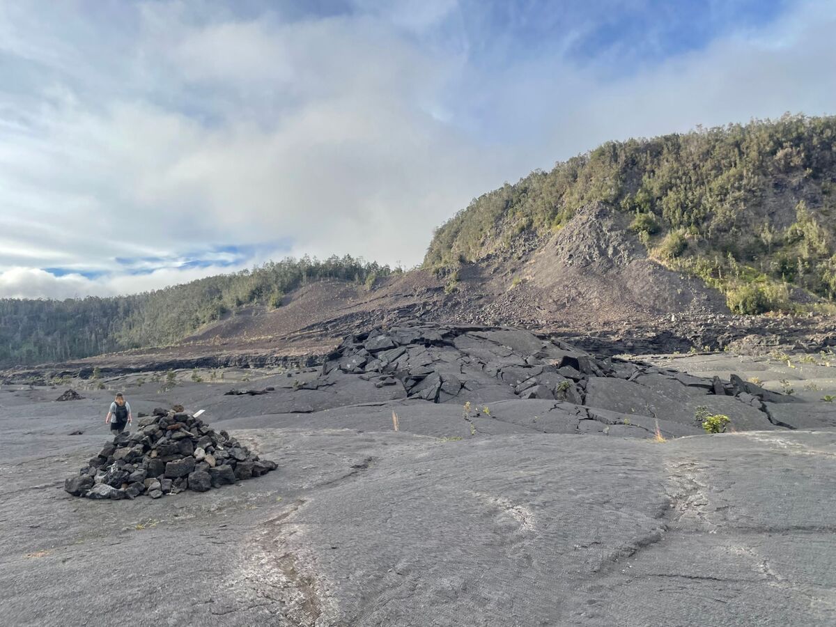 A hiker passing a stone cairn on the cracked lava floor of Kīlauea Iki, with the crater’s forested wall behind