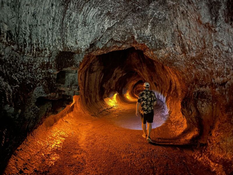 A visitor walking through the lit, rust-orange passage of Nāhuku (Thurston Lava Tube)
