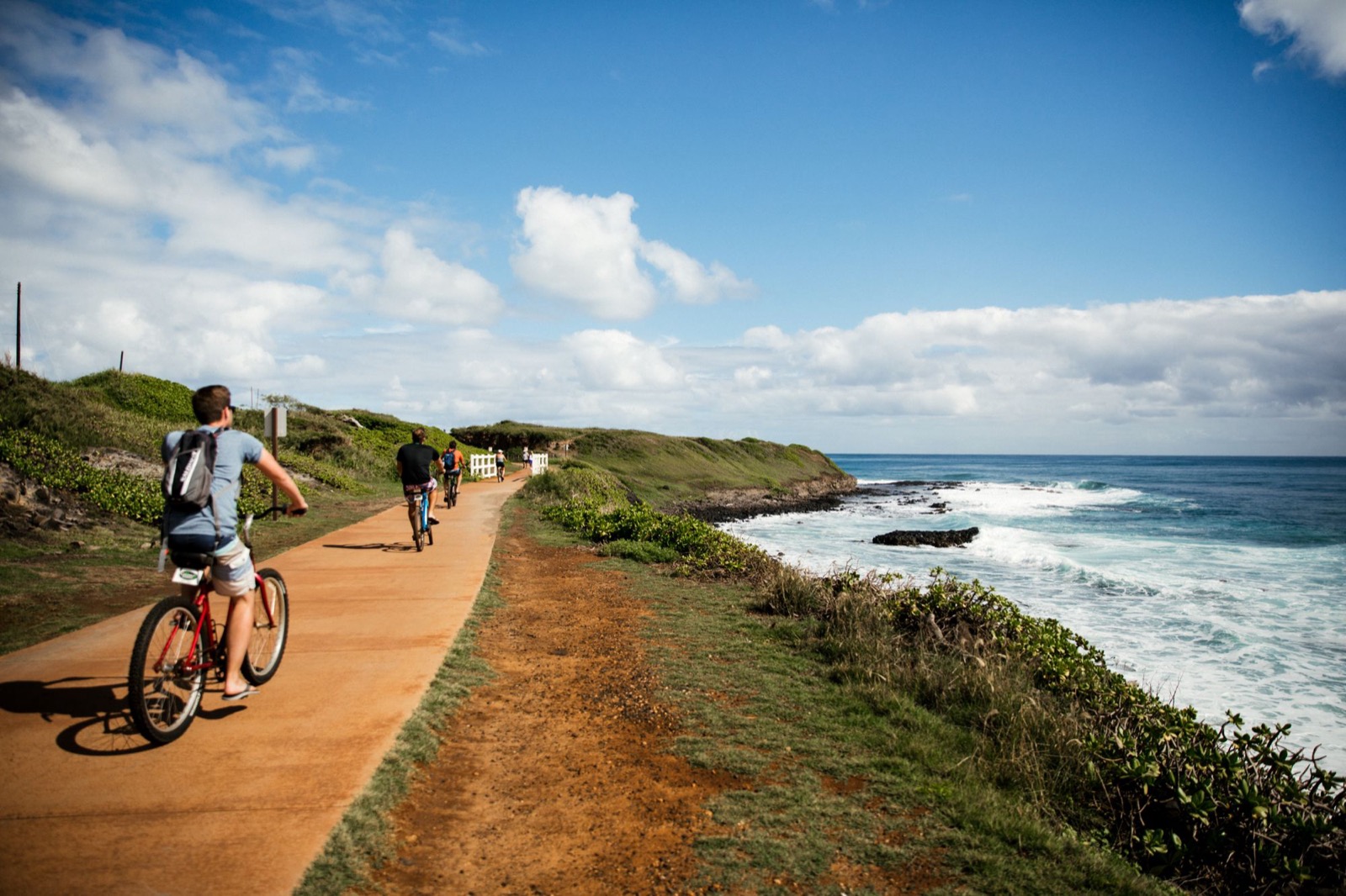 Cyclists riding a coastal path above surf breaking on a rocky shoreline under a blue sky