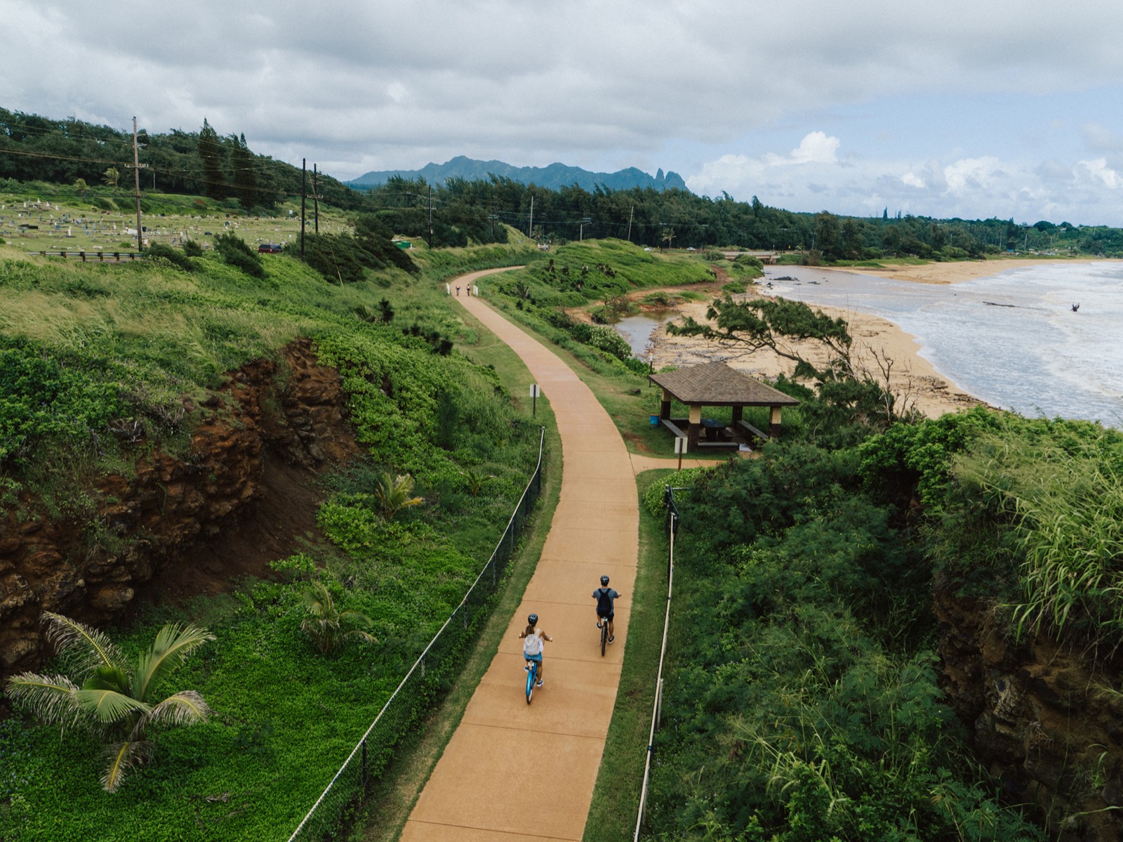 Aerial view of two cyclists on the coastal path, between a green hillside and a beach, with a small pavilion on the shore