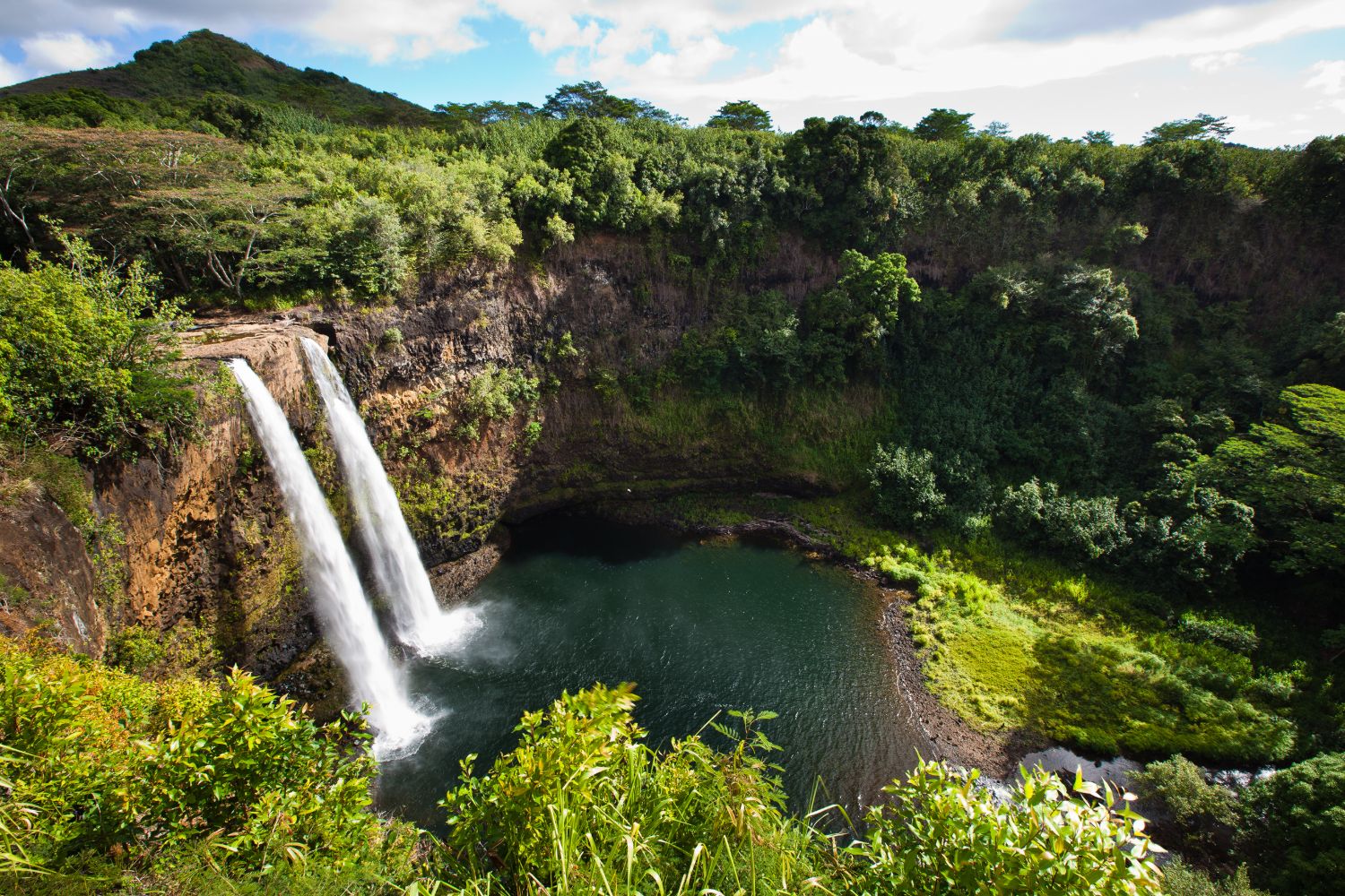 Twin streams of Wailua Falls dropping into a green pool below a forested ridge