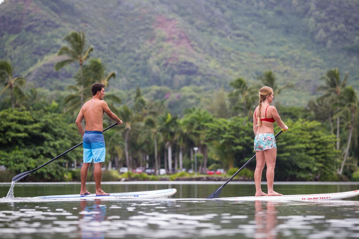 Two stand-up paddleboarders on the calm Wailua River below palm trees and green hills