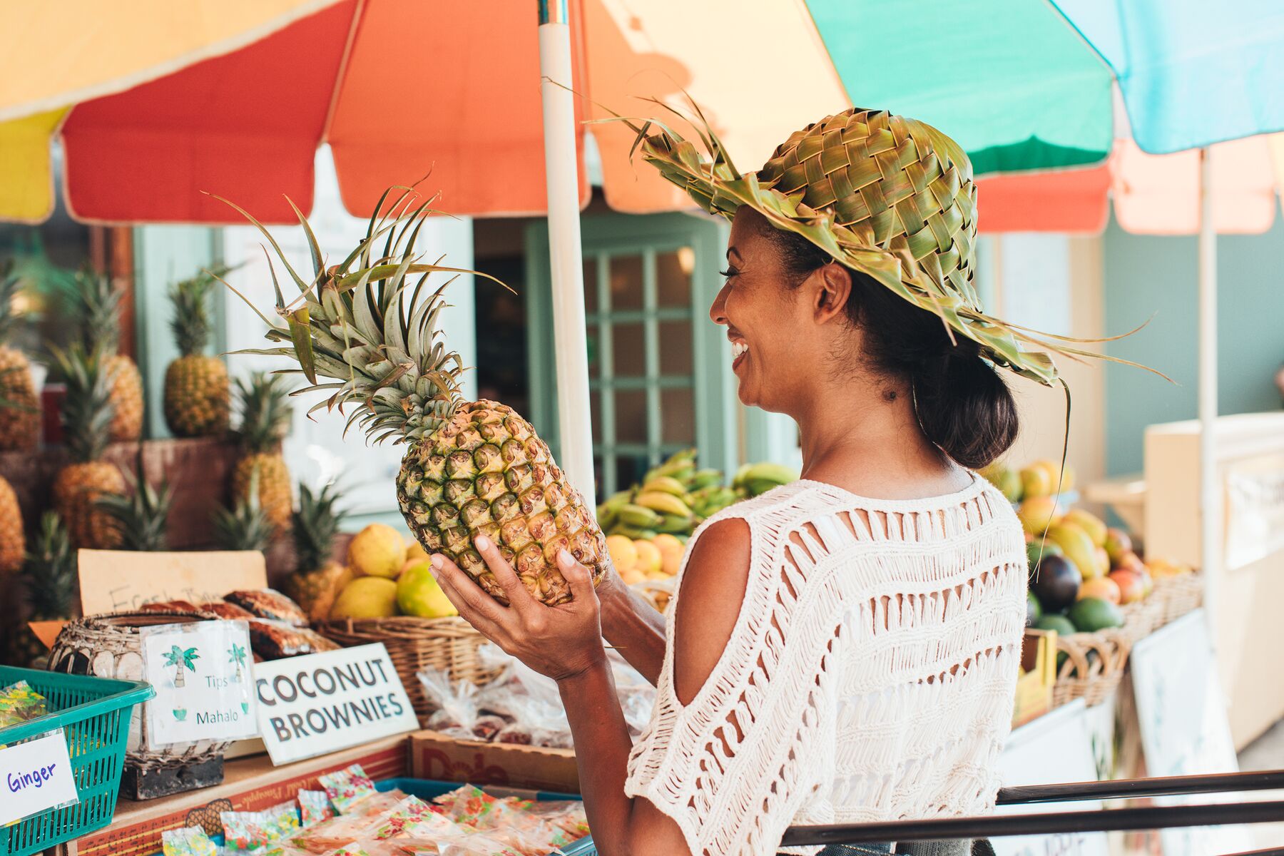 A smiling woman in a palm-frond hat holding a large pineapple at a farmers-market stall with a colorful umbrella