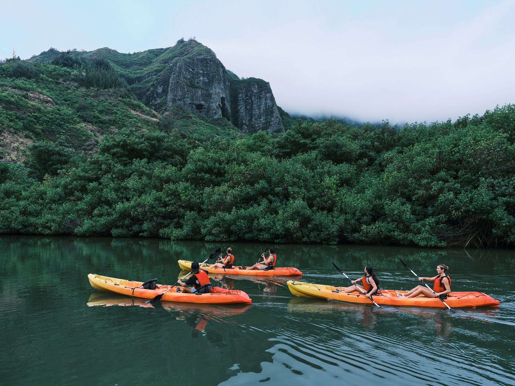 Two orange tandem kayaks on the still, dark water of Hulēʻia Stream, with a misty green peak behind
