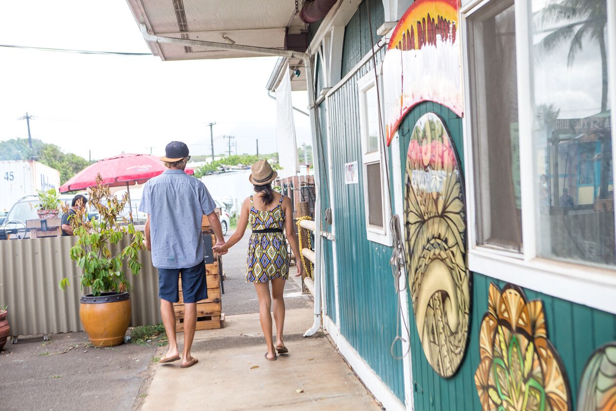 A couple walking past a teal wooden building with round murals, small shops and a red market umbrella