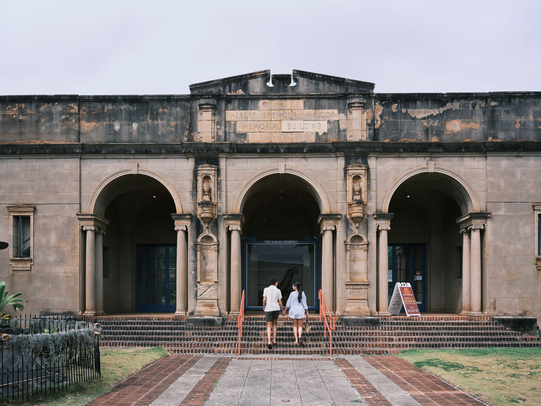 The stone front of the Kauaʻi Museum in Līhuʻe, with three arched entries and two visitors walking up the steps