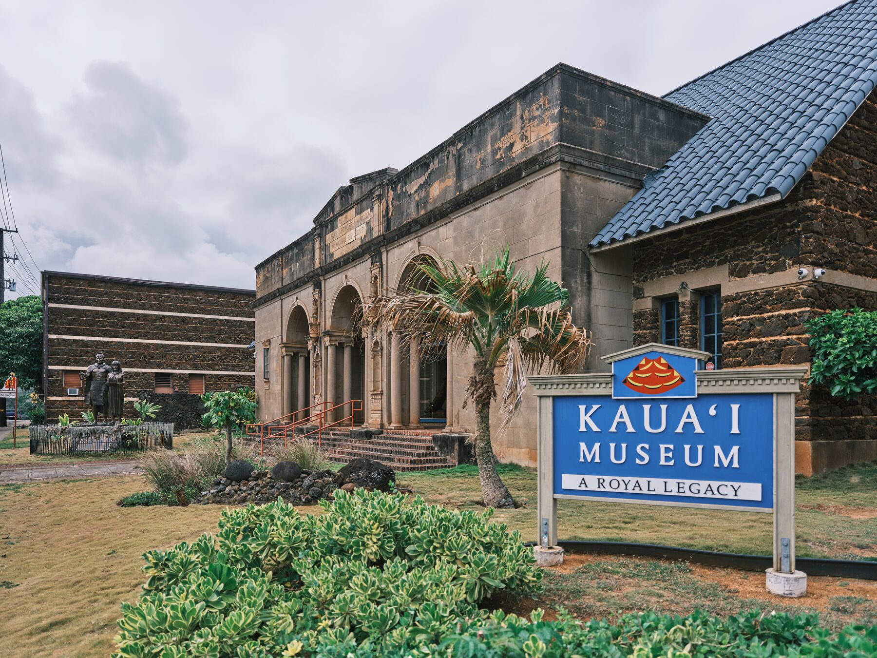 The blue “Kauaʻi Museum – A Royal Legacy” sign in front of the museum’s dark stone arches and blue roof, under an overcast sky
