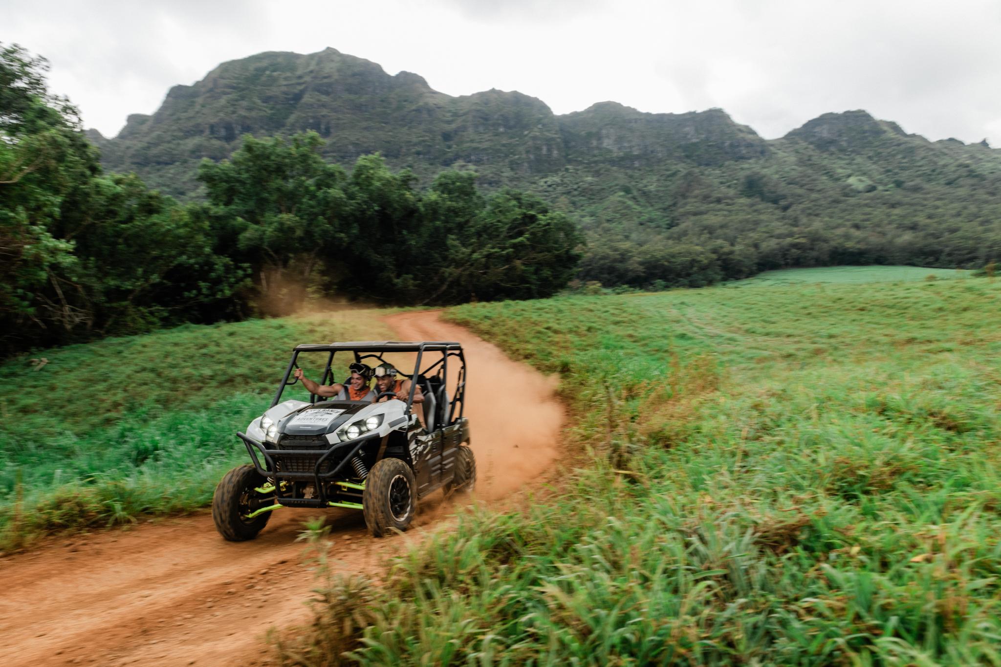 Two people laughing in an ATV kicking up red dust on a dirt track through green pasture, with a steep forested mountain ridge behind
