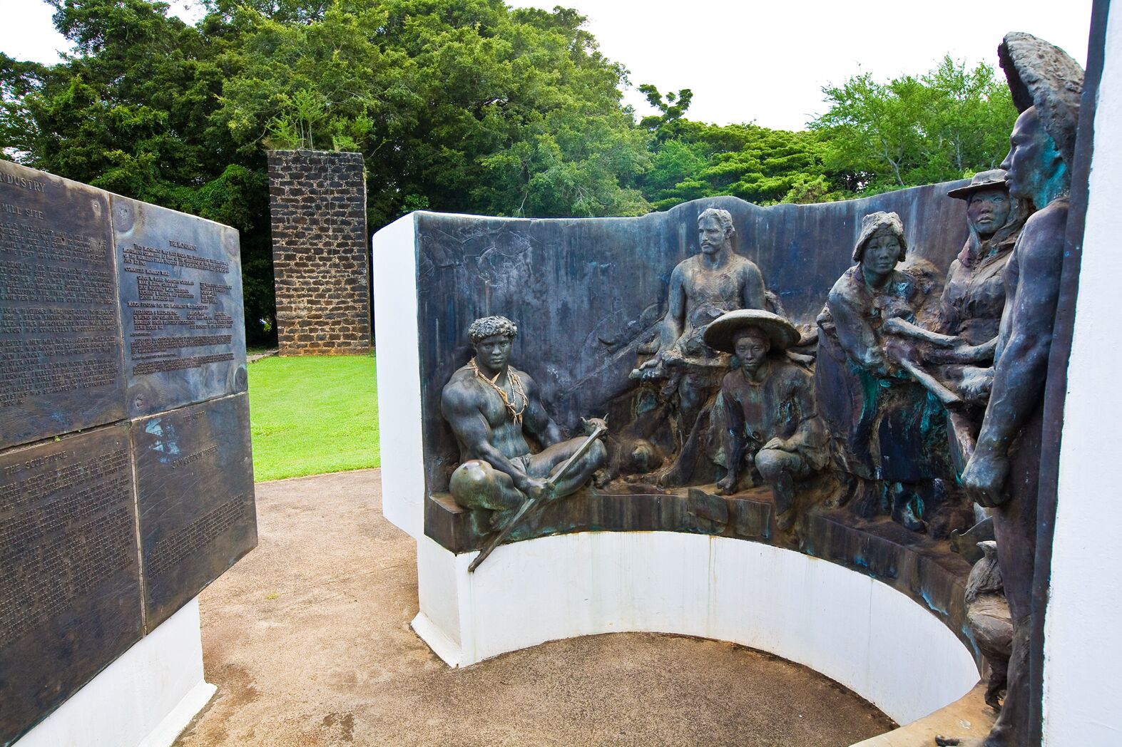 A bronze sculpture of several figures in period dress before a curved white wall, with a tall plaque to one side and a sugar-mill chimney in the background
