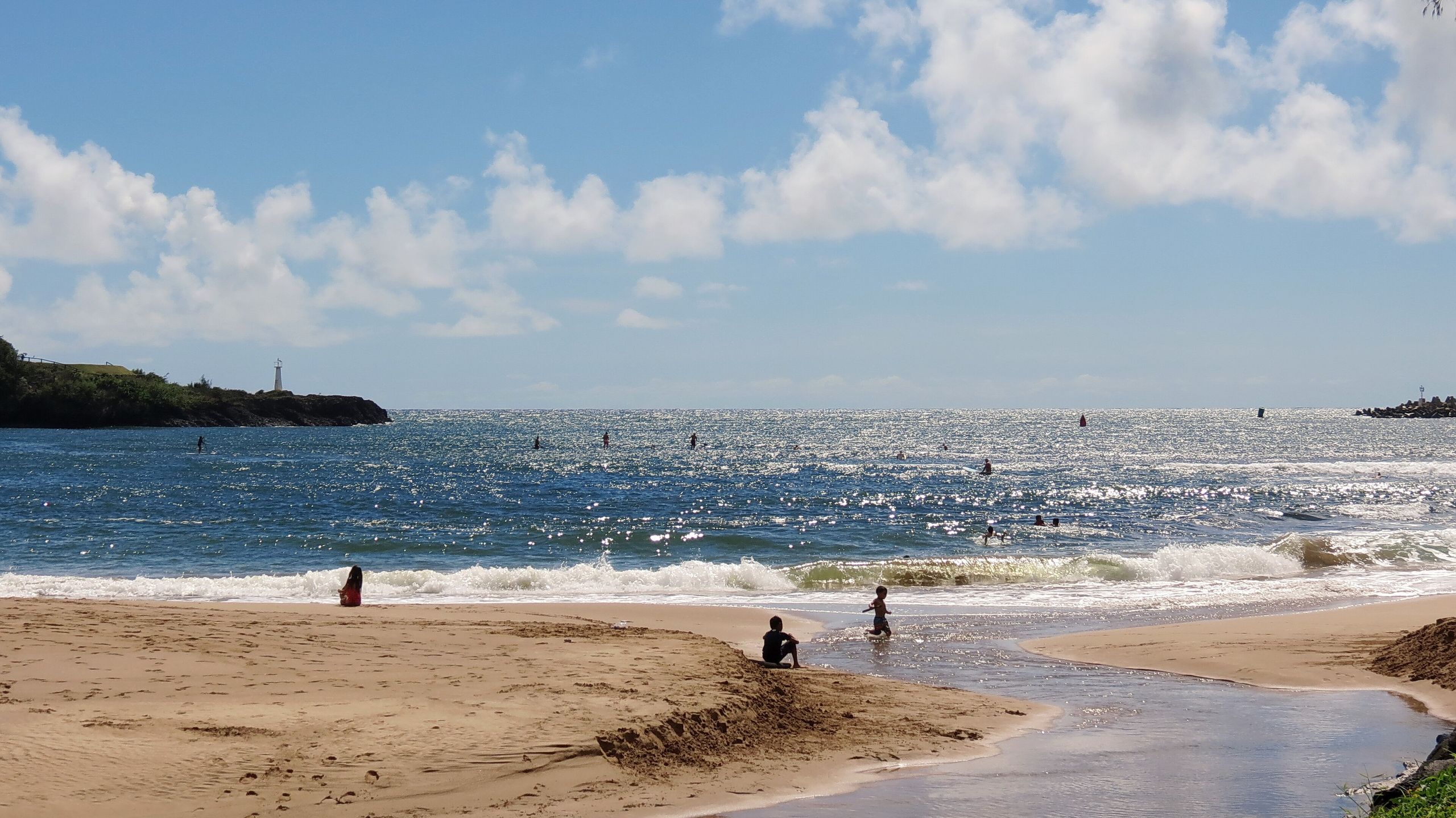 Nāwiliwili Beach Park: a small stream crosses the sand into the bay, with paddleboarders offshore and the Ninini Point lighthouse on the headland at left