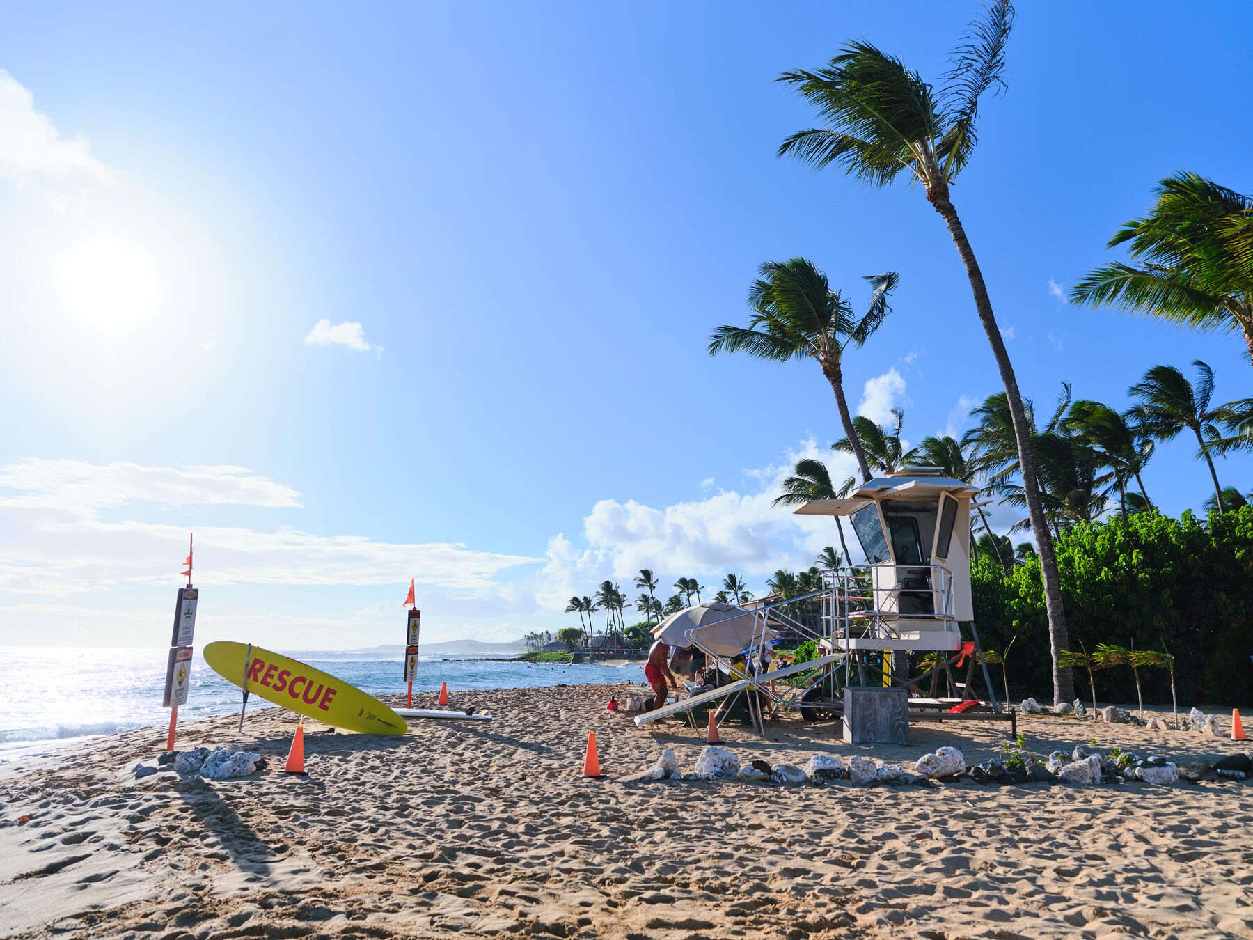 A tan lifeguard tower and a yellow “RESCUE” board on a sunny beach, with palm trees blowing on the right