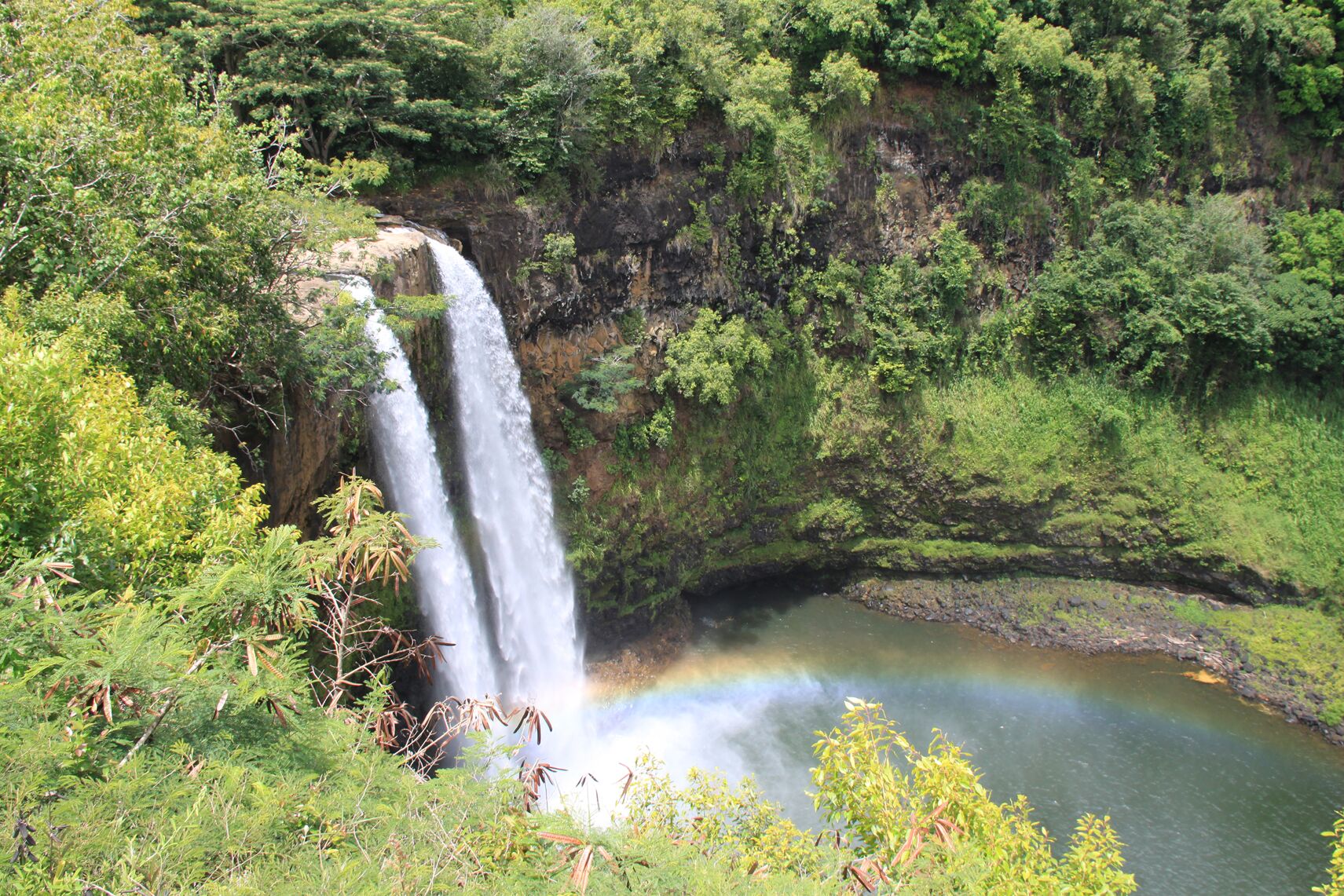Wailua Falls, twin streams dropping down a tall green cliff into a pool, with a faint rainbow in the mist