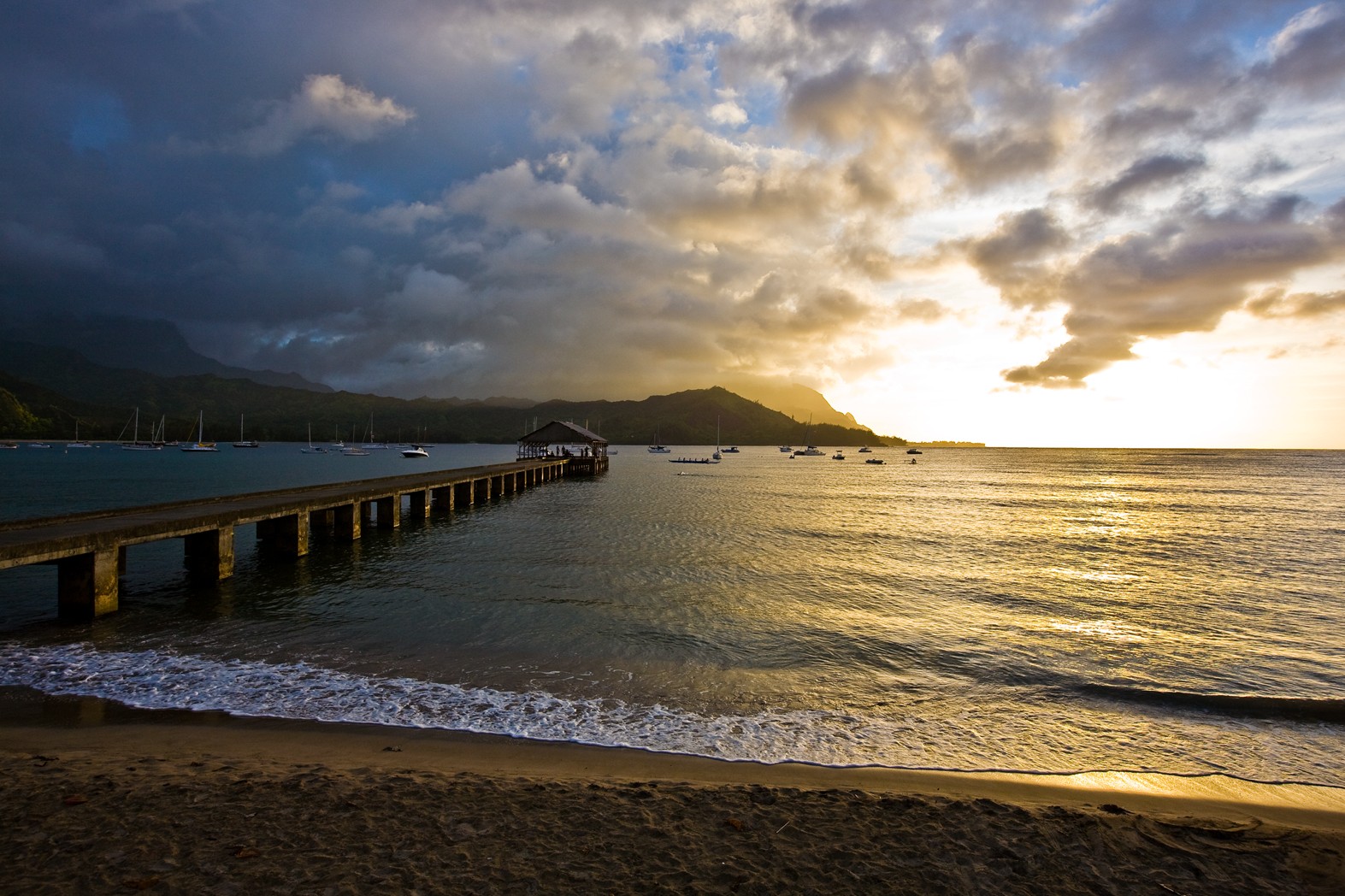 Golden sunset light on Hanalei Bay beside Hanalei Pier, sailboats moored below storm clouds