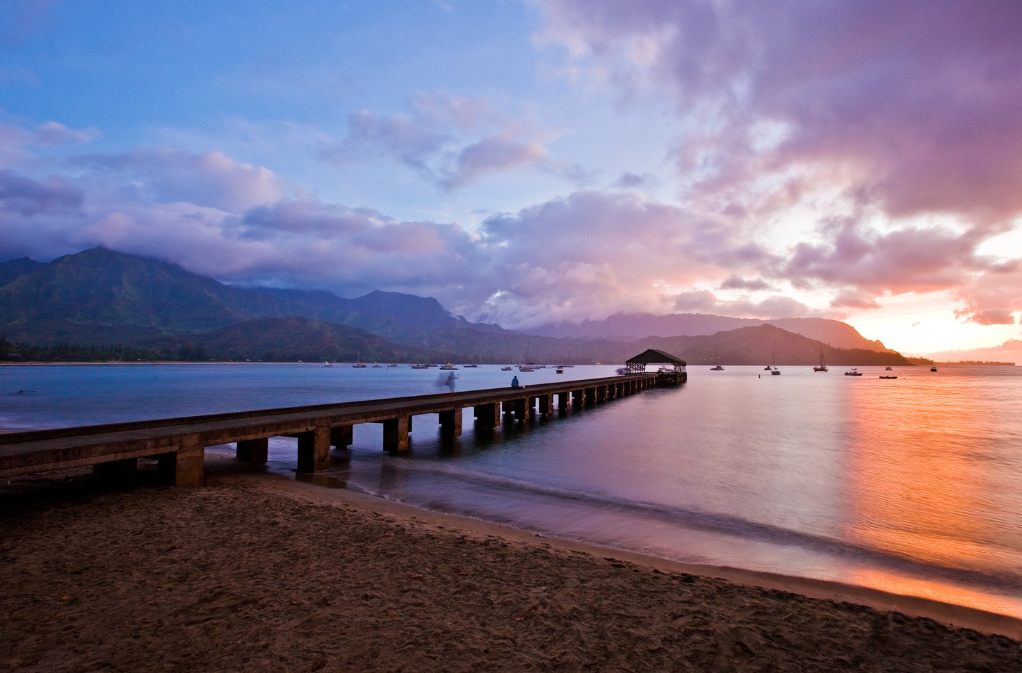 Hanalei Pier reaching into Hanalei Bay at sunset, moored sailboats and cloud-topped mountains behind