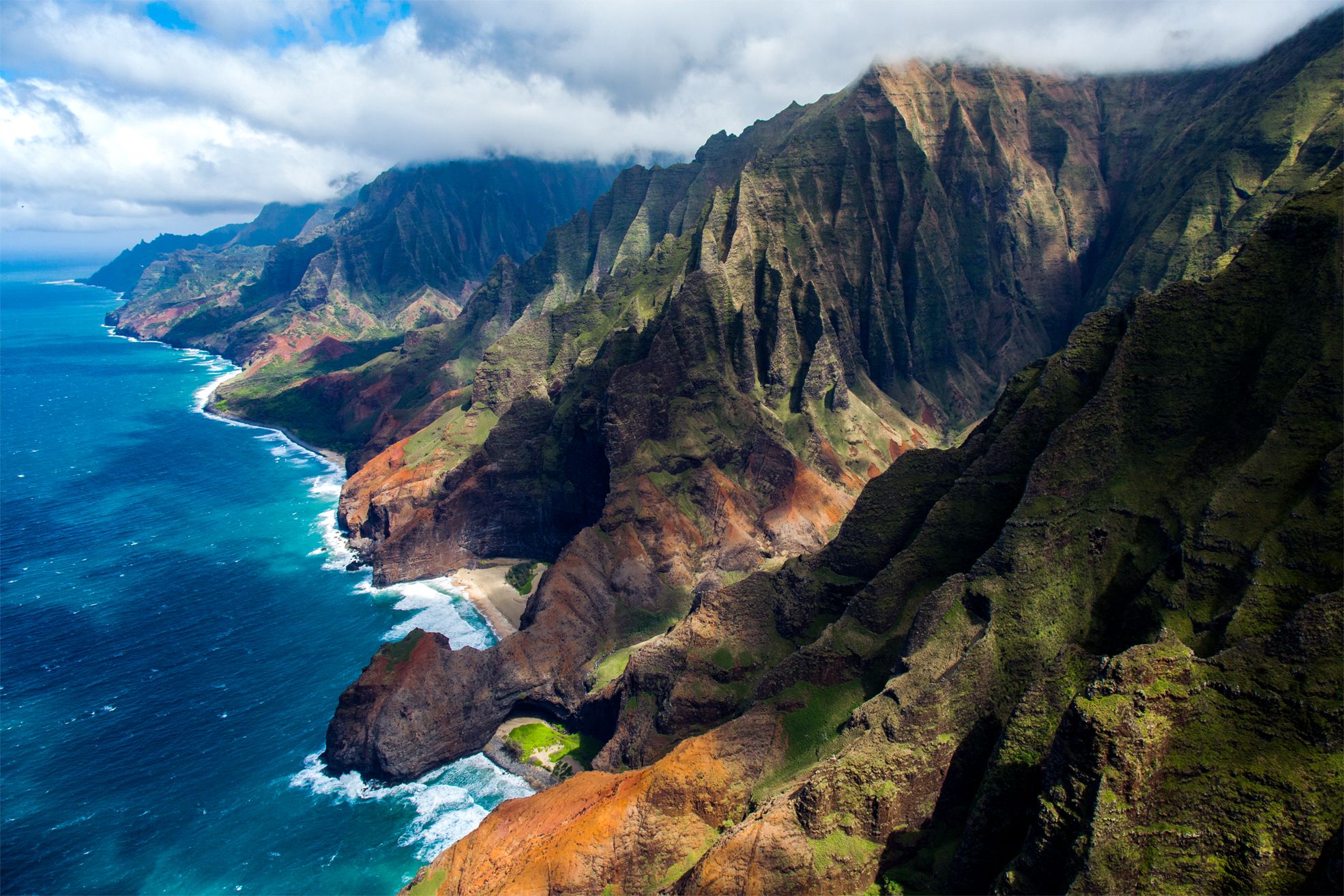 Nāpali Coast ridges and sea cliffs above surf-lined coves, clouds on the peaks