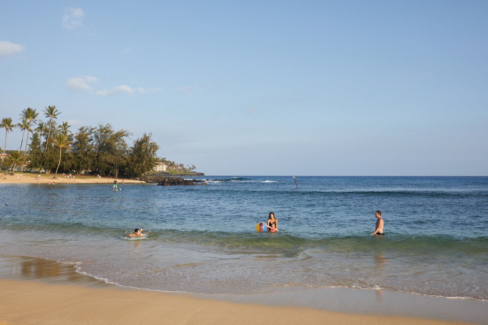 A family swimming and playing with a beach ball in the gentle surf at Poʻipū Beach, with palms on the point behind