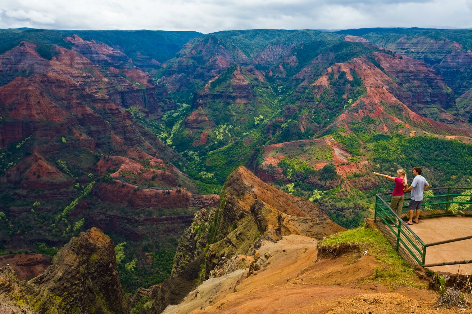 A woman and a man stand at a green railing on a paved lookout, the woman pointing across the deep, multicolored gorge of Waimea Canyon under an overcast sky
