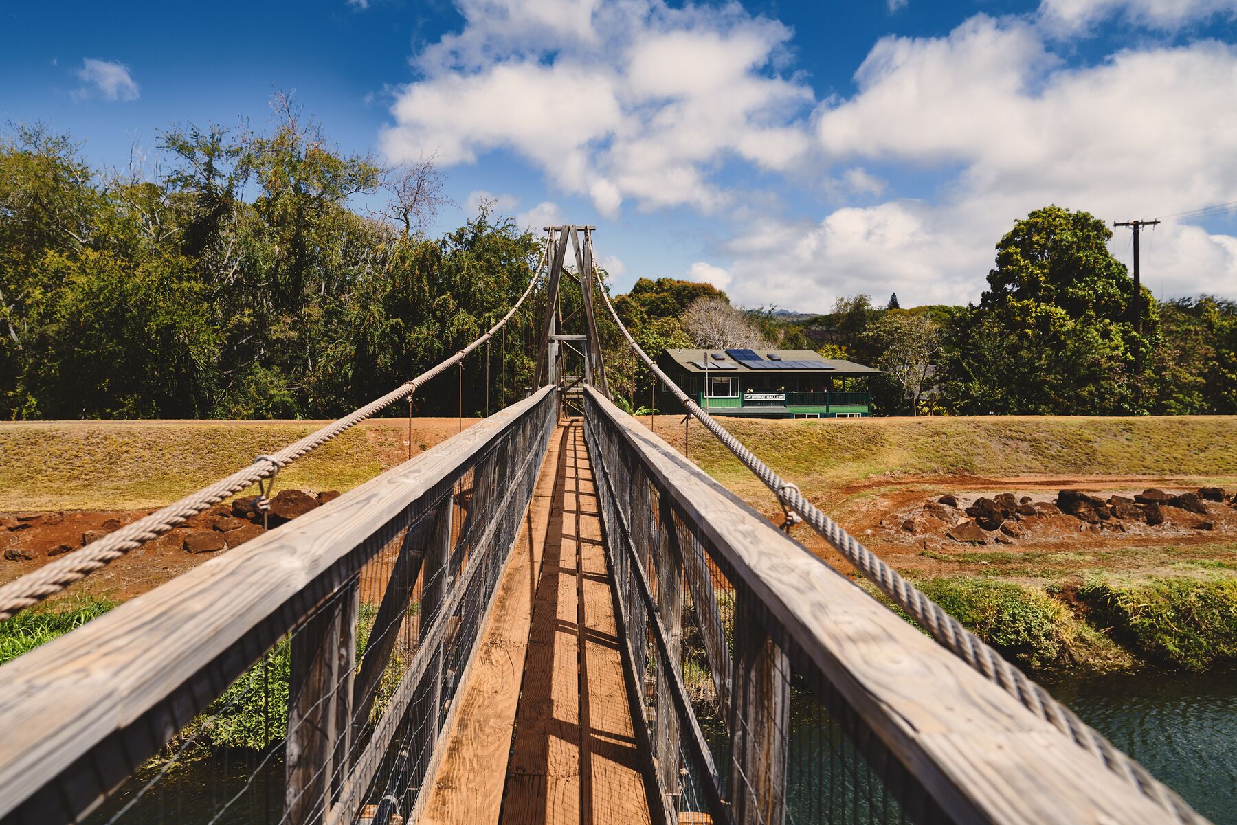 Looking along the wooden deck of Hanapēpē’s suspension footbridge, with cable railings leading to the far bank and trees under a blue sky