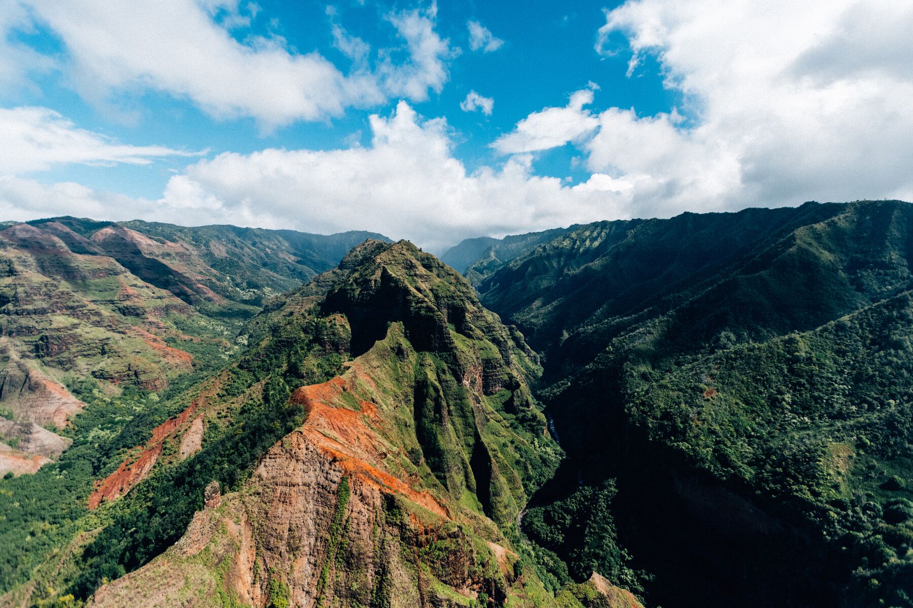 A tour helicopter, small against the scene, flies past a sharp green and red ridge in Waimea Canyon, with clouds above