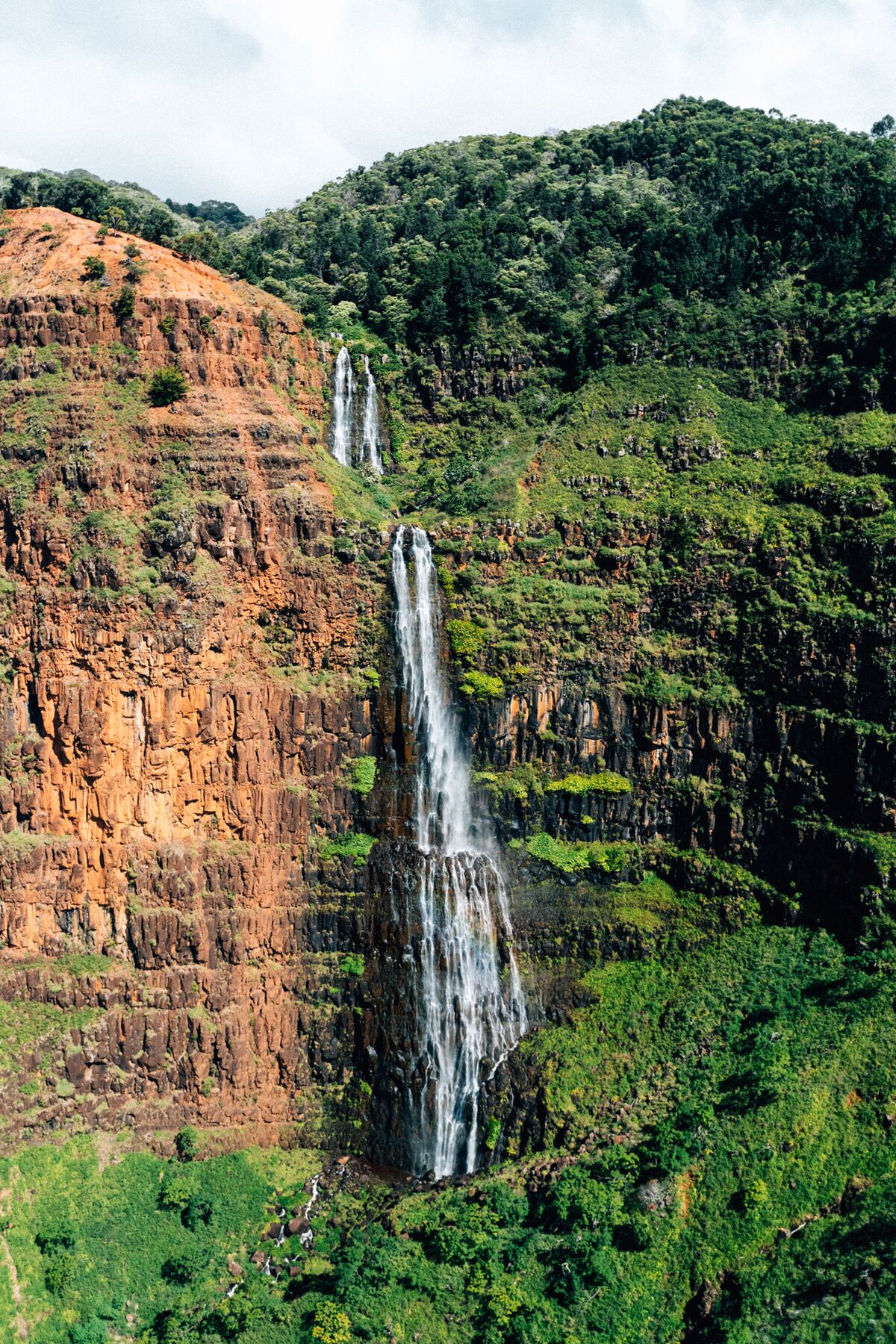 A tour helicopter flies past a tall, narrow waterfall pouring down a red, fern-covered cliff in Waimea Canyon