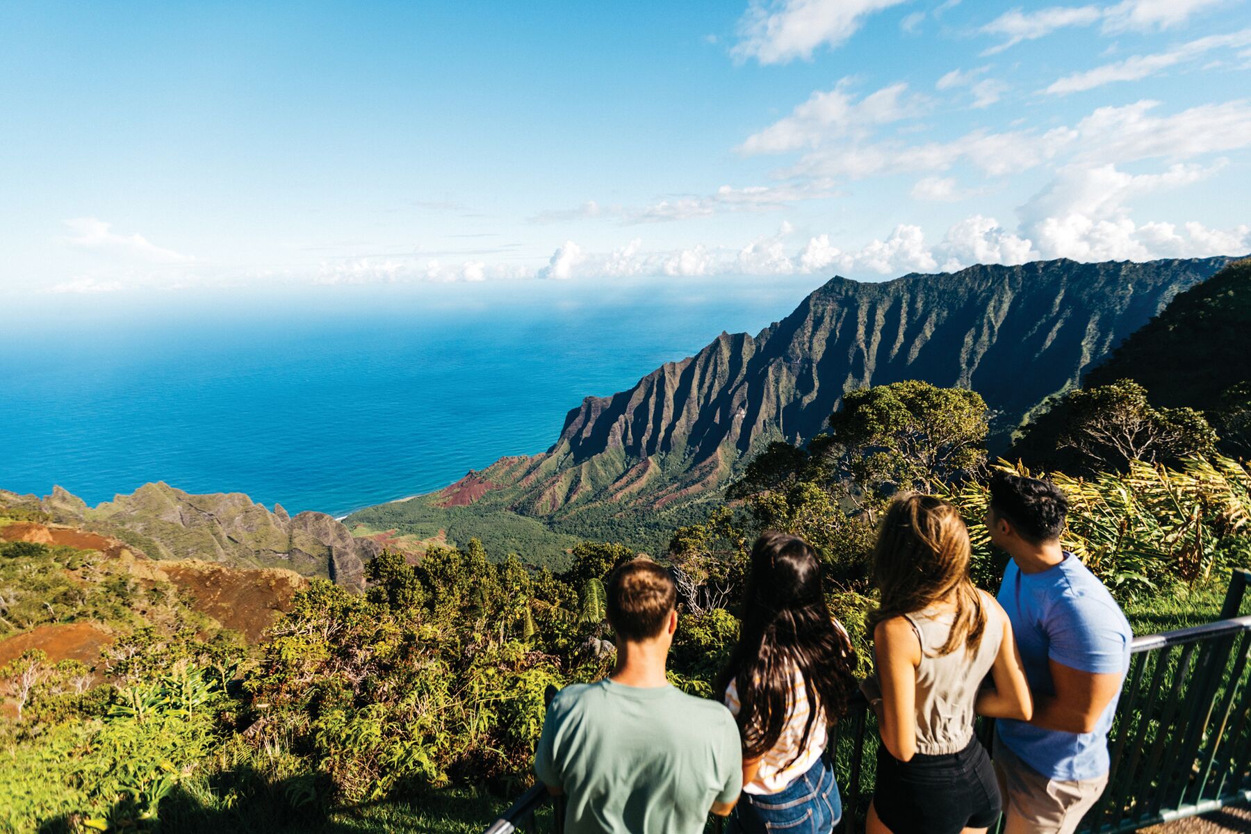 Four young visitors stand at the Kalalau Lookout, looking out at the green Nāpali ridges and the ocean below