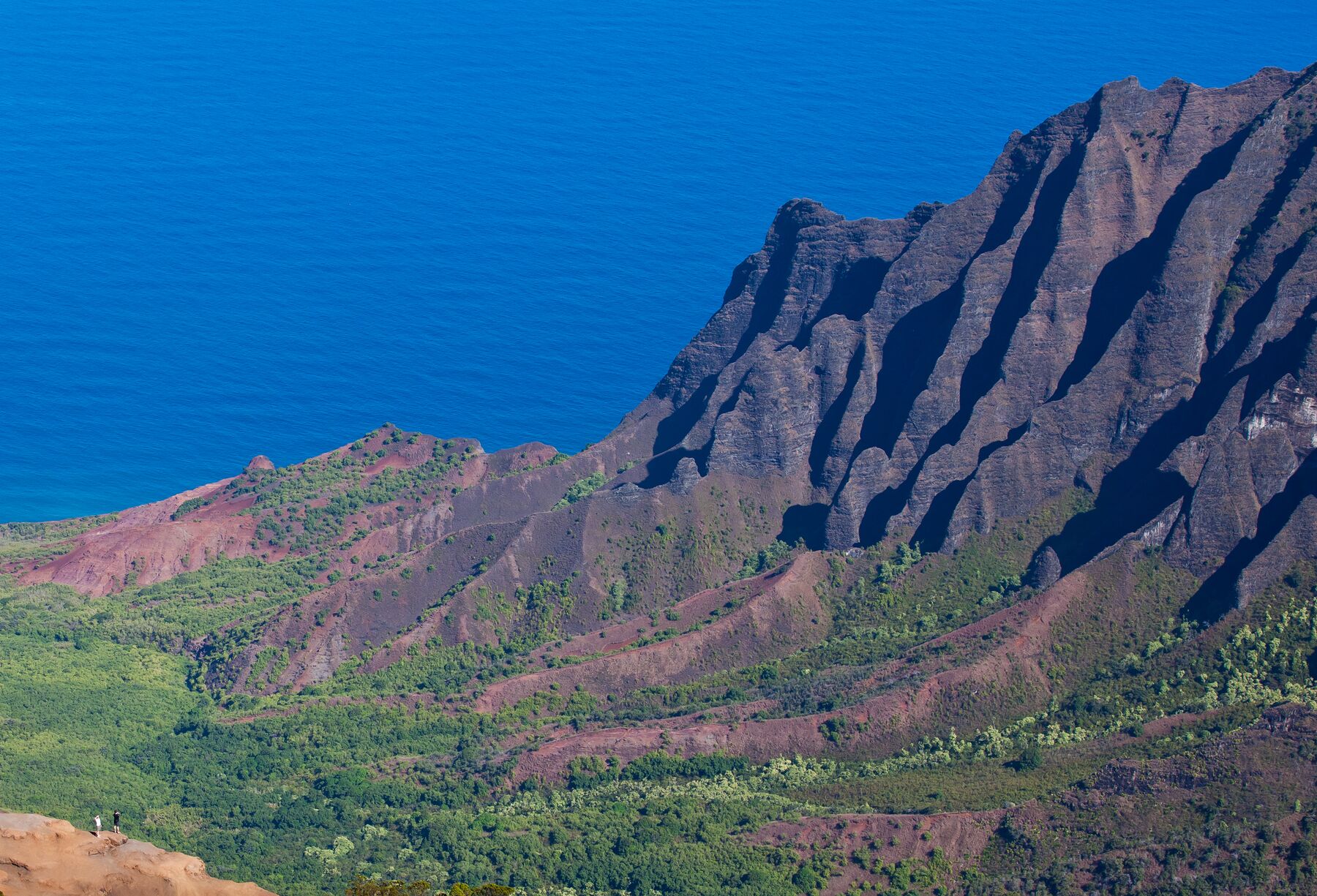 The steep, deeply fluted brown and green cliffs of Kalalau Valley on the right, with the blue Pacific beyond and two tiny figures on a rocky outcrop at lower left