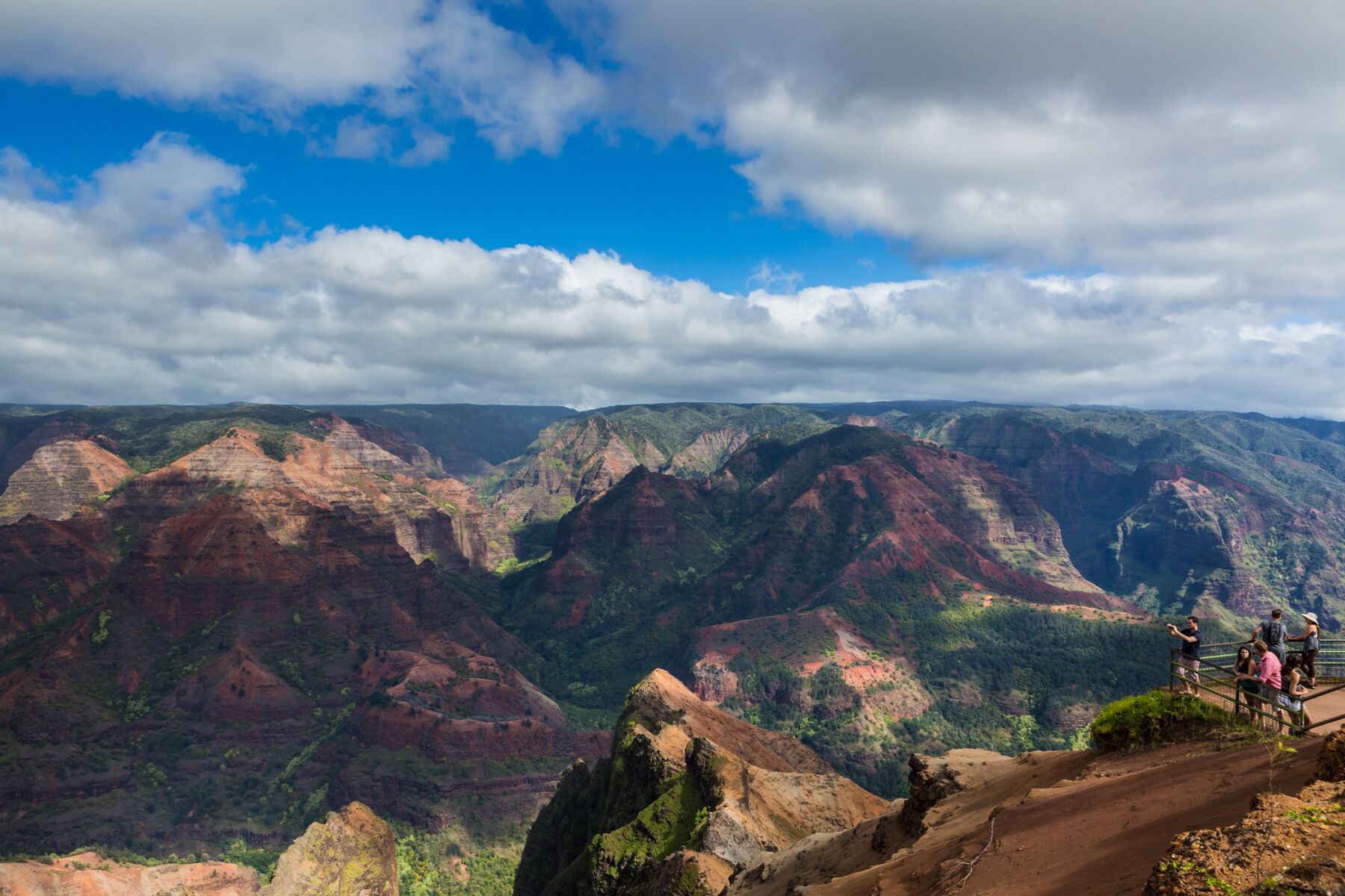 A small group of visitors stands behind a wooden post-and-rail fence on a red-dirt viewpoint, looking out over Waimea Canyon’s red and brown ridges under a partly cloudy sky